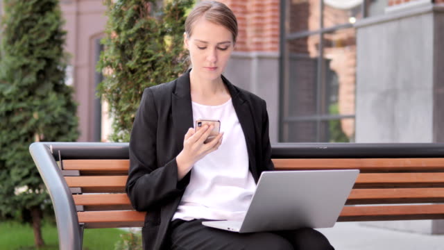 Junge-Geschäftsfrau-nutzt-Smartphone-und-Laptop,-sitzt-auf-Bank