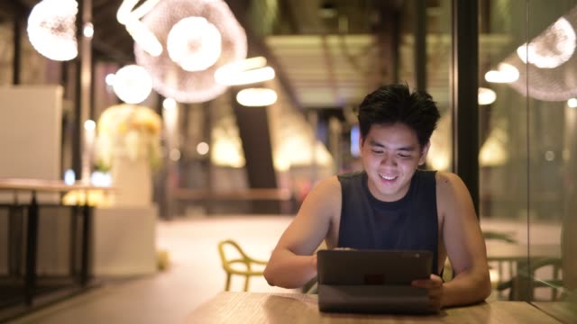 Junge-glückliche-stattliche-asiatische-Mann-mit-digitalen-Tablet-im-Restaurant-in-der-Nacht
