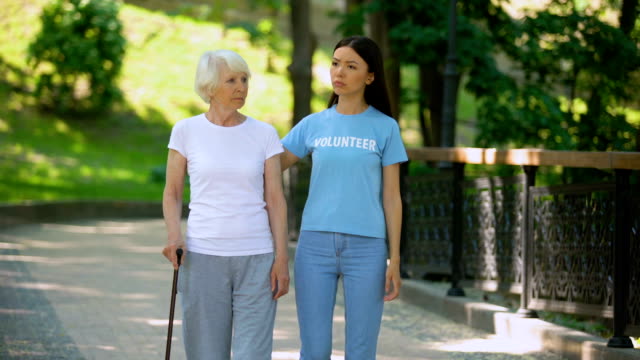 Depressed-mature-woman-with-walking-stick-and-female-volunteer-in-garden,-rehab