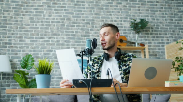 Joyful-guy-reading-in-microphone-holding-paper-making-podcast-in-studio