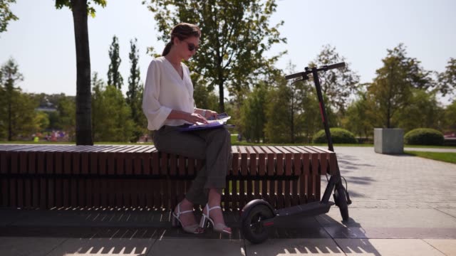 Young-and-independent-businesswoman-on-a-bench-in-a-park,-next-to-an-electric-scooter.