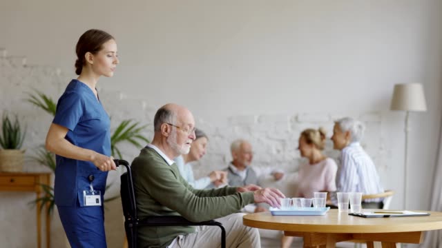 Female-caregiver-pushing-wheelchair-with-disabled-elderly-man.-Senior-taking-pills-and-glass-of-water-on-table-and-joining-friends-talking-on-sofa-in-nursing-home