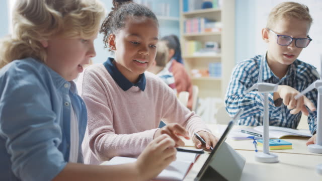 In-der-Grundschule:-Klasse-intelligenter-Kleiner-Kinder-arbeitet-als-Team-mit-Tablet-Computern,-um-Windkraftanlagen-zu-programmieren.-Klassenzimmer-mit-Kindern-Lernen-über-umweltfreundliche-Formen-erneuerbarer-Energie