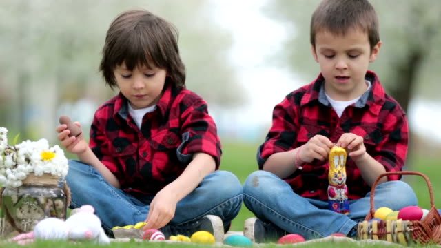 Two-children,-boy-brothers,-eating-chocolate-bunnies-and-having-fun-with-easter-eggs-in-the-park,-beautiful-spring-blooming-garden