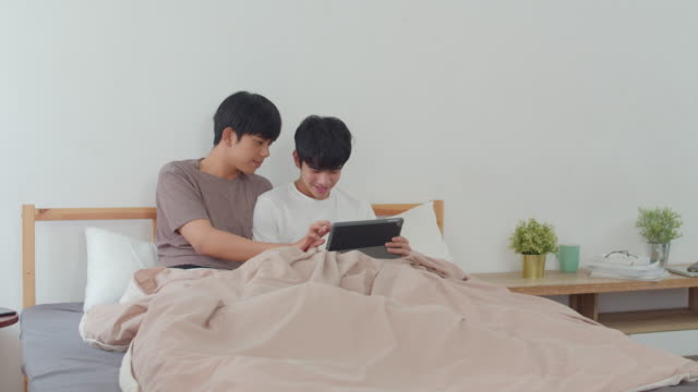 Pareja-gay-asiática-usando-tableta-en-casa.-Jóvenes-hombres-LGBTQ-asiáticos-feliz-descanso-juntos-después-de-despertar,-revisar-el-correo-y-las-redes-sociales-acostados-en-la-cama-en-el-dormitorio-en-casa-en-el-concepto-de-la-mañana.