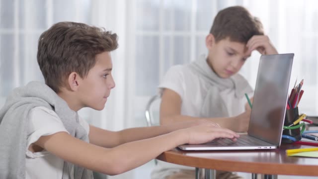 Seitenansicht-Nahaufnahme-von-kaukasischen-Jungen-spielen-Online-Spiele-auf-Laptop.-Sein-Zwillingsbruder-sitzt-im-Hintergrund-und-macht-Hausaufgaben.-Unterschiedliche-Einstellung-zum-Studium,-Bildungskonzept,-Internetsucht.