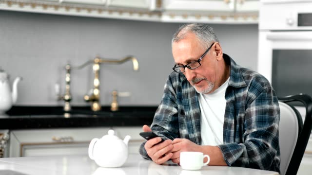 Smiling-mature-bearded-man-in-glasses-chatting-using-smartphone-enjoying-coffee-break