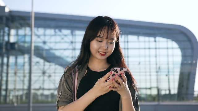 Angenehme-unbeschwerte-junge-asiatische-Frau-tippt-SMS-auf-ihrem-Handy-in-der-Nähe-des-Flughafengebäudes