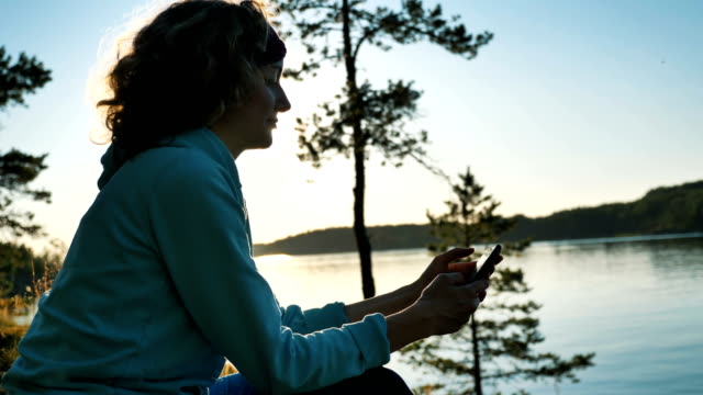 young-woman-uses-smartphone-sitting-on-bank-of-tranquil-lake