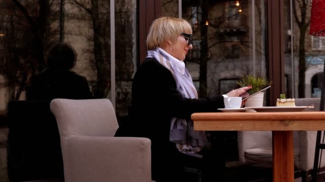 Una-mujer-elegante-con-unas-gafas-de-sol-está-sentada-en-una-cafetería-con-una-tableta-y-café.