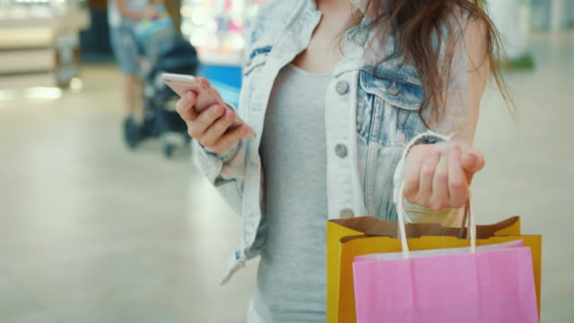Ein-Mädchen-geht-durch-ein-Einkaufszentrum-und-sms-auf-ihrem-Smartphone.-Sie-trägt-Einkaufstaschen.-Nahaufnahmen.-4K