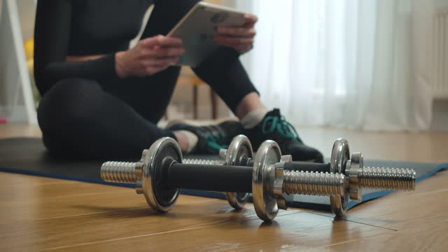 Close-up-of-dumbbells-with-blurred-slim-sportswoman-surfing-Internet-on-tablet-at-the-background.-Unrecognizable-young-Caucasian-woman-having-break-during-training-at-home.-Cinema-4k-ProRes-HQ.