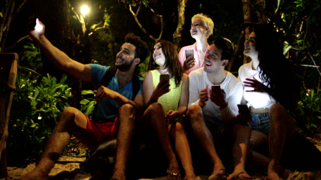 La-gente-toma-selfie-sentado-en-el-banco-en-el-parque-de-la-noche-usando-el-teléfono-celular-inteligente,-amigos-charlando-en-línea-al-aire-libre