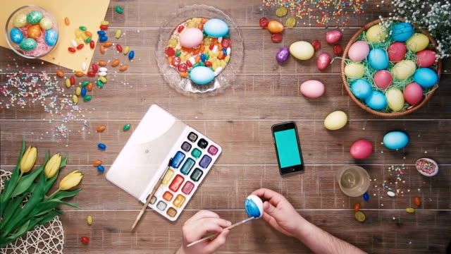 Man-painting-easter-egg-and-smart-phone-with-green-screen-lies-on-table-decorated-with-easter-eggs.-Top-view