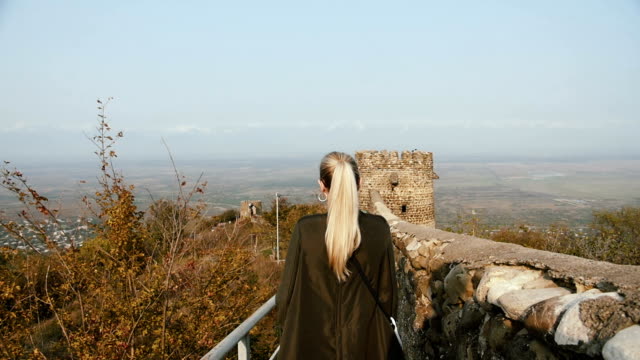 A-young-woman-tourist-in-a-green-leather-jacket-walks-the-fortress-wall-of-an-ancient-castle.-Shot-in-slow-motion-100fps