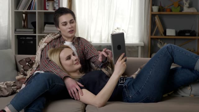 Lesbian-couple-is-resting-on-the-couch,-using-tablet-computer,-scrolling-photos-on-tablet-60-fps