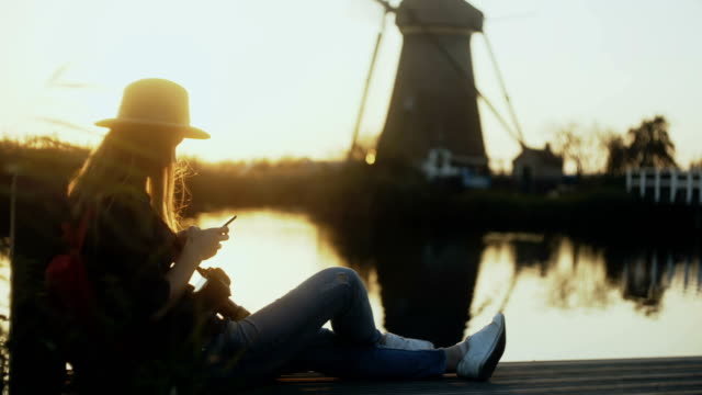 Mädchen-sitzt-mit-Smartphone-auf-Sonnenuntergang-See-Kai.-Frau-mit-mobile-app-im-Freien.-Traditionelle-holländische-Windmühle-Landschaft.-4K