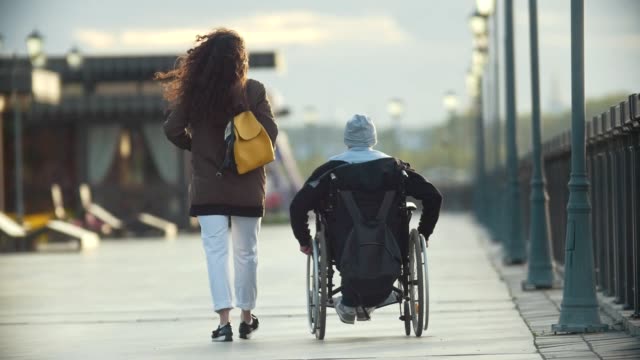Mujer-joven-feliz-con-un-hombre-discapacitado-en-silla-de-ruedas-caminar-juntos-en-el-muelle