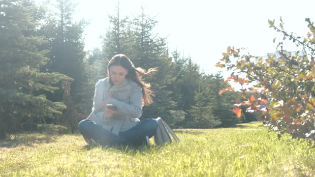 Young-beautiful-brunette-woman-sits-on-grass-on-strong-wind-in-spring-park-and-types-a-message-in-mobile-phone.