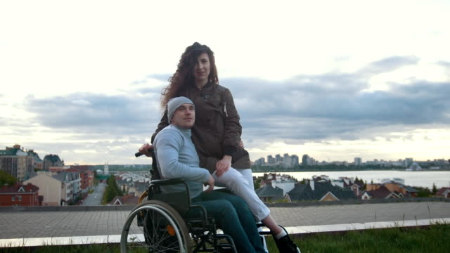 Portrait-of-a-happy-disabled-man-in-a-wheelchair-embraces-with-young-woman-outdoors