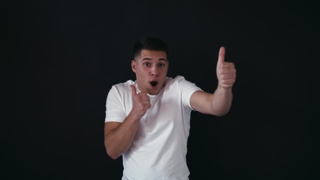 Funny-guy-stands-and-poses-on-camera.-Young-man-keeps-his-big-thumbs-up-on-hands.-Guy-has-playful-mood.-He-poses.-Isolated-on-black-background