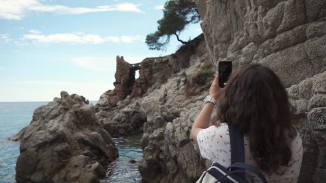 Brunette-woman-is-taking-photos-of-beautiful-rocks-on-seashore-by-mobile-phone
