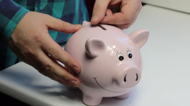On-the-table-there-is-a-piggy-bank-in-the-form-of-a-pink-pig.-The-man-in-the-hands-of-the-coin,-which-he-puts-in-turn-in-a-piggy-bank.