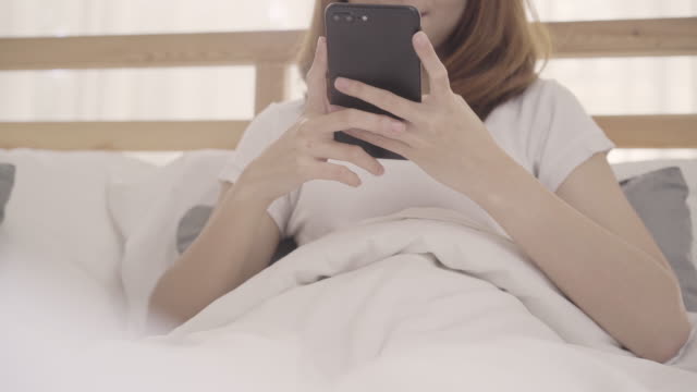 Junge-Asiatin-mit-Smartphone-auf-Bett-liegend,-nach-dem-Aufwachen-am-Morgen,-schöne-attraktive-Japanerin-lächelnd-im-Schlafzimmer-zu-Hause-entspannen.-Genießen-Zeit-Lifestyle-Frauen-bei-Wohnkonzept.