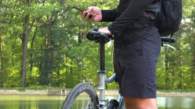 A-cyclist-stands-with-a-bike-on-a-shore-by-a-lake-and-works-on-a-smartphone---closeup