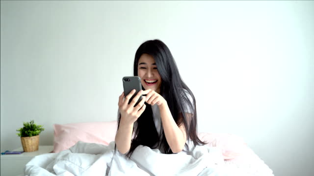 Una-hermosa-mujer-asiática-sentada-en-una-cama-está-usando-un-teléfono-móvil-para-hablar-con-las-redes-sociales-con-amigos.