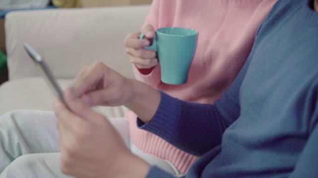 Young-Asian-couple-using-tablet-and-drinking-coffee-in-living-room-at-home,-couple-enjoy-love-moment-while-lying-on-sofa-when-relaxed-at-home.-Enjoying-time-lifestyle-family-at-home-concept.