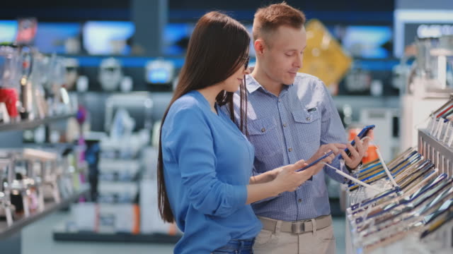 Man-holding-smart-phone-and-pointing-on-other-phone-which-is-in-woman-hands.-Tech-store-interior