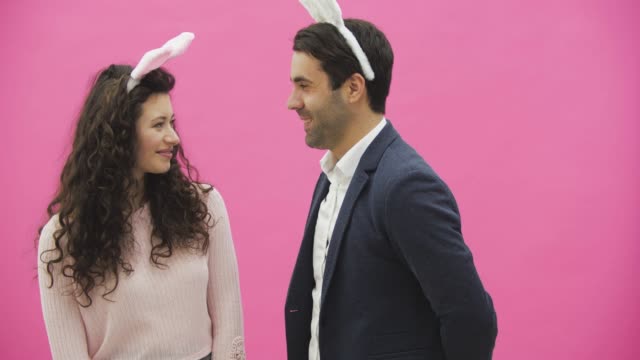 Young-sexy-couple-on-pink-background.-With-hackneyed-ears-on-the-head.-During-this-man-gives-a-soft-toy-with-a-hare-to-his-wife.-Having-kissed-looking-at-the-camera.