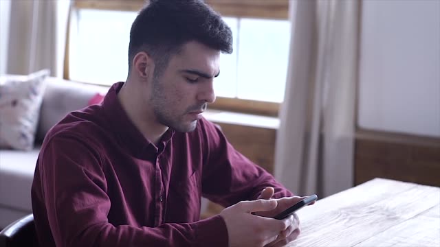 Happy-Young-Man-Uses-Smartphone-while-Sitting-on-a-chair-at-Home.-Man-Browses-Through-Internet,-Watches-Videos-and-Uses-Social-Networks-at-Home.