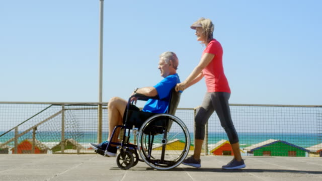 Side-view-of-active-senior-Caucasian-woman-pushing-senior-man-on-wheelchair-at-beach-4k