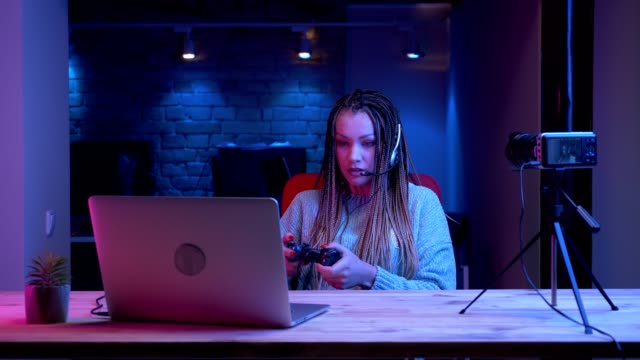 Closeup-shoot-of-young-attractive-female-blogger-with-dreadlocks-in-headphones-playing-video-games-on-the-laptop-and-cheerfully-talking-while-streaming-with-the-neon-background-indoors