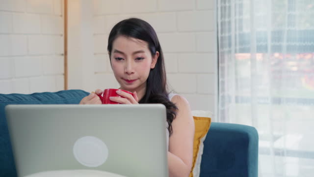 Junge-freiberufliche-asiatische-Frau,-die-am-Laptop-arbeitet-und-soziale-Medien-prüft-und-Kaffee-trinkt,-während-sie-auf-dem-Sofa-liegen,-während-sie-sich-im-Wohnzimmer-zu-Hause-entspannen.-Lifestyle-Frauen-bei-Hauskonzept.