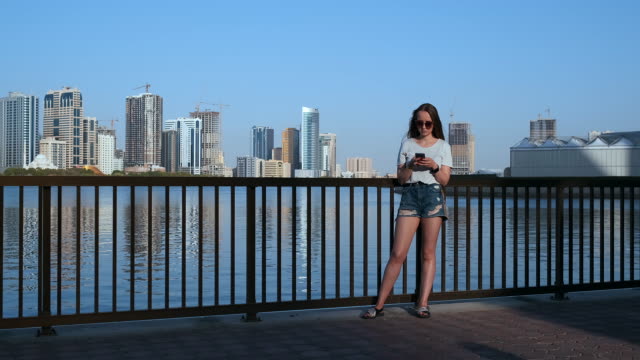 Beautiful-girl-with-long-hair-in-sunglasses-using-smartphone-app-at-sunset-river-quay-near-4K.