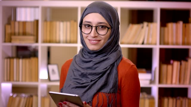 Sesión-de-primer-plano-de-joven-atractiva-estudiante-musulmana-en-hiyab-usando-la-tableta-y-mirando-la-cámara-sonriendo-de-pie-en-el-interior-de-la-biblioteca