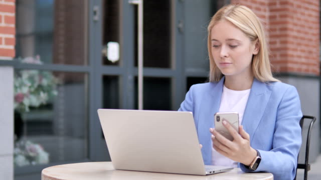 Young-Businesswoman-Using-Smartphone-and-Laptop,-Sitting-Outdoor
