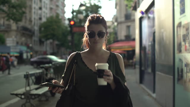 Atractiva-mujer-caucásica-con-gafas-de-sol,-pecas,-piercings-y-pelo-rojo-desplazando-las-redes-sociales-en-su-teléfono-inteligente-y-bebiendo-café-caminando-por-la-calle,-durante-el-verano-soleado-en-París.-4K.