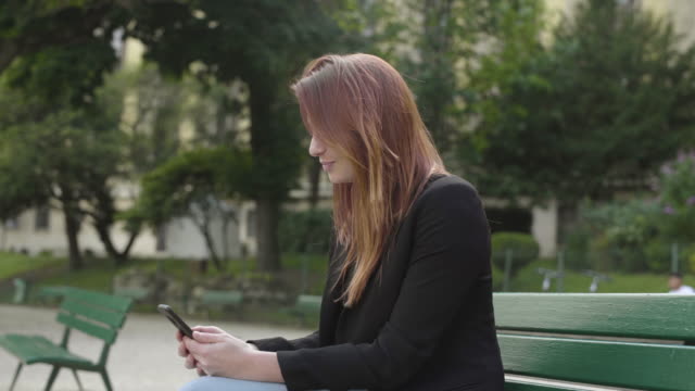 Smiling-Businesswoman-using-smartphone-Sitting-on-bench-in-park-in-Paris,-texting-with-mobile-telephone-outside,-typing-text-message-on-cell-phone.-Content,-Videos,-Red-hair.-Social-media-outdoor.
