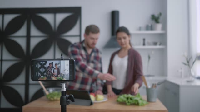 Vegan-Blog,-Vlogger-Kerl-und-Mädchen-bereiten-gesundes-Essen-mit-Gemüse-und-Grüns-in-der-Küche,-während-Kamera-Smartphone-zeichnet-Video-für-Follower-in-sozialen-Netzwerken