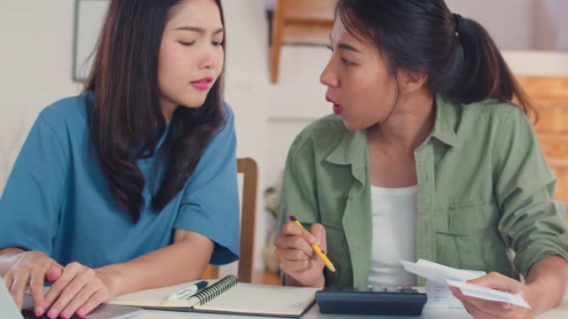 Pareja-lesbiana-asiática-de-negocios-usando-presupuesto-récord-calculadora,-impuestos,-documento-financiero-en-computadora-portátil-trabajando-en-la-cocina.