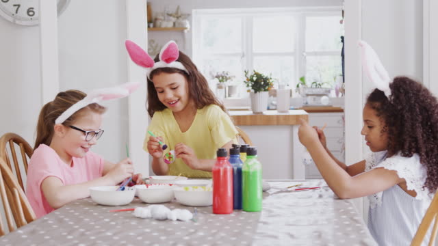 Grupo-de-chicas-sentadas-alrededor-de-la-mesa-de-la-cocina-con-orejas-de-conejo-decorando-huevos-para-Semana-Santa---tiro-en-cámara-lenta