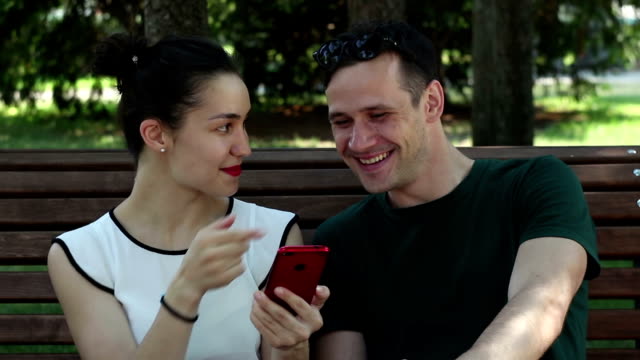 Una-joven-pareja-amorosa,-en-un-día-soleado,-está-discutiendo-activamente-un-nuevo-teléfono-inteligente-mientras-se-sienta-en-un-banco-en-el-parque.