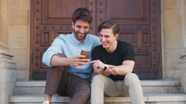 Männliche-Homosexuell-Paar-sitzen-im-Freien-auf-Stufen-des-Gebäudes-Blick-auf-Handy