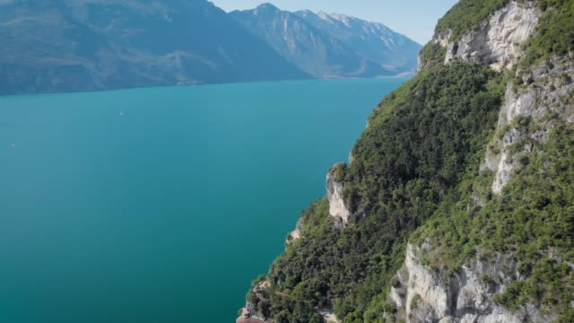 Drohne-fliegt-über-erstaunlichen-Bergsee-mit-türkisfarbenem-Wasser,-entlang-hohen-Berg-bedeckten-Wald-in-sonnigen-Tag