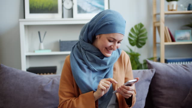Hermosa-mujer-de-Oriente-Medio-en-hiyab-usando-teléfono-inteligente-en-casa-sonriendo