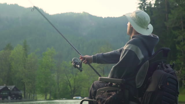 Cámara-lenta-de-pescador-discapacitado-en-silla-de-ruedas-eléctrica-pescando-en-un-hermoso-lago-cerca-del-bosque-y-la-montaña-en-la-parte-posterior,-en-la-puesta-de-sol